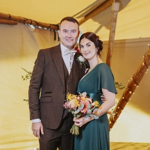 A couple dressed formally stands together inside a warmly lit tent decorated with wooden beams and string lights.