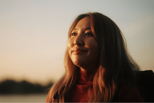 A woman with long hair smiling softly while looking into the distance during sunset, with a calm outdoor background.-1