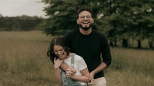 A couple laughing together in an open field, the man holding the woman from behind as they both smile broadly