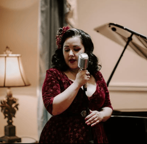 Becki singing into a vintage-style microphone in a red velvet dress, in front of a piano