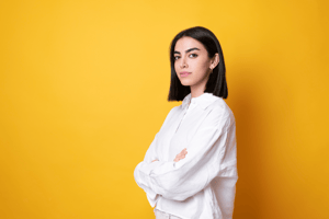 Dark haired young woman in a white shirt standing in front of a  yellow background with her arms crossed 